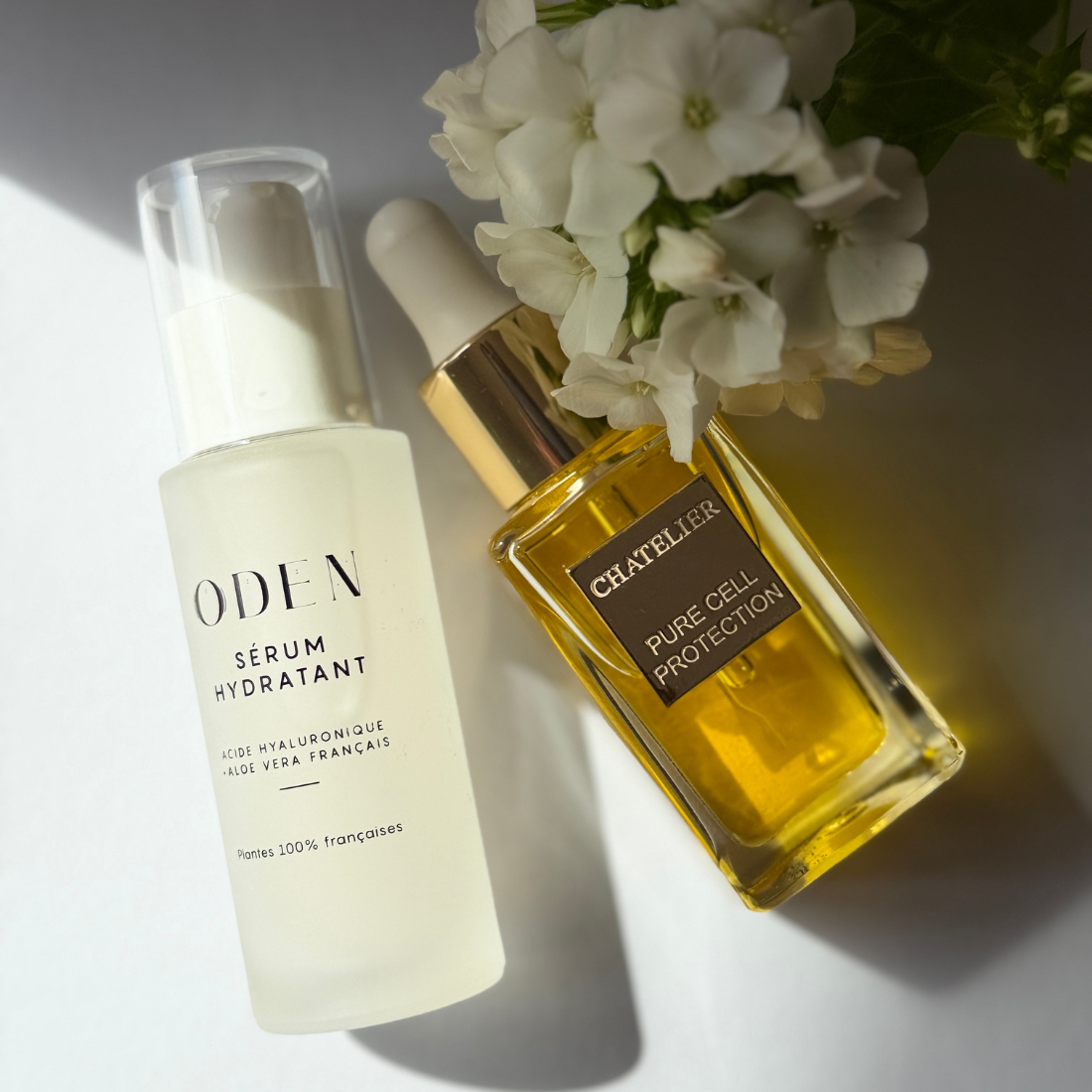 Two skincare bottles arranged on a white surface with white flowers: a frosted white bottle labeled ODEN Serum Hydratant and a gold-accented amber bottle labeled CHATELIER Pure Cell Protection, photographed in bright directional light with strong shadows.