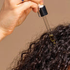 Hand applying a dropper of oil to curly hair against a beige background